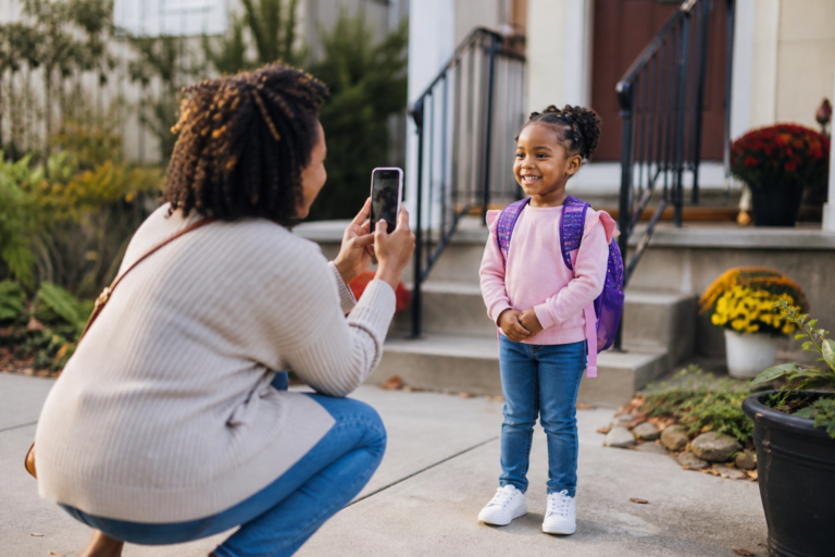 Black mother taking a photo of preschool child with a backpack on a sidewalk