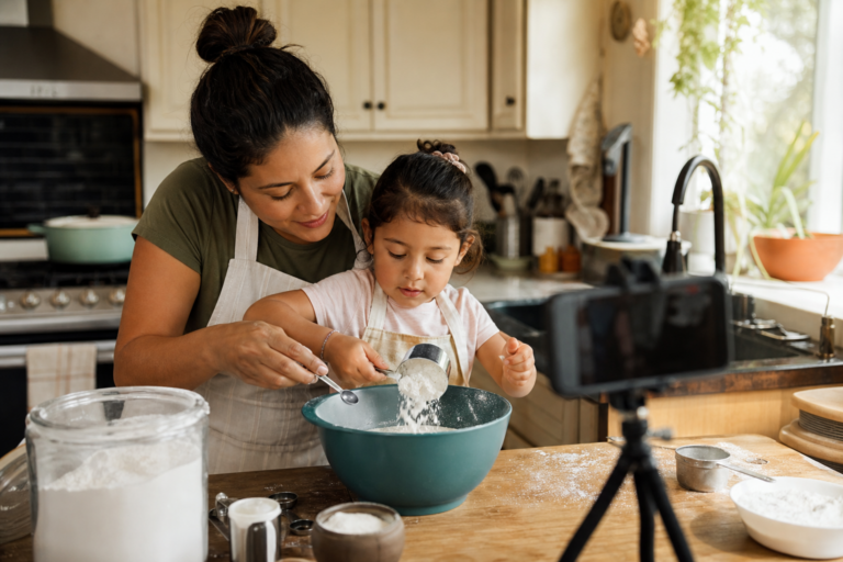Latina mother and young child baking together in a family kitchen while a phone records nearby